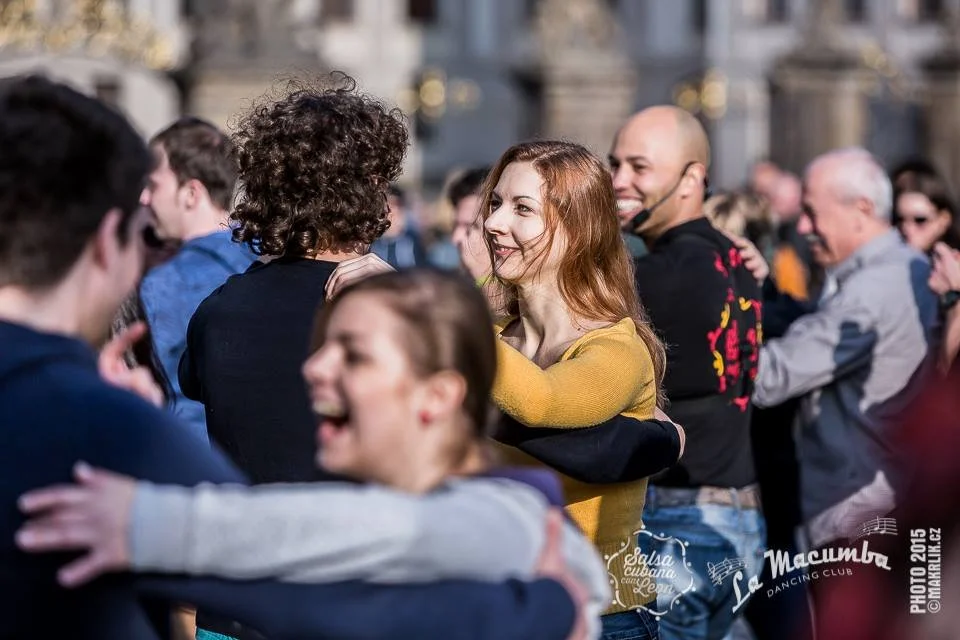 People dancing and socializing outdoors, smiling, with some hugging each other, in a lively crowd during daytime.