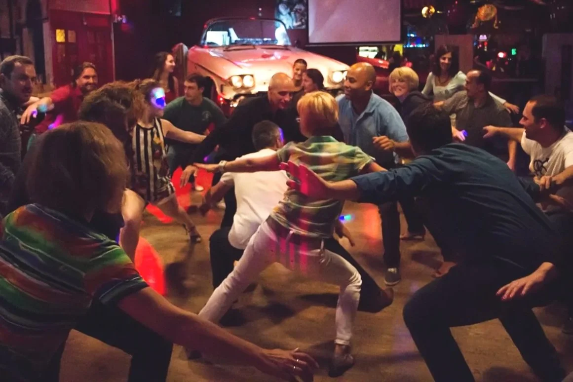 People dancing energetically in a dimly lit bar or club with retro cars in the background.