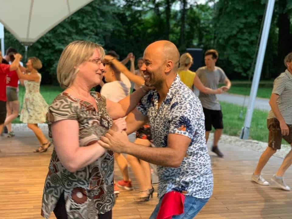 A man and woman are dancing and smiling at each other outdoors during a party or gathering, with other couples dancing in the background.