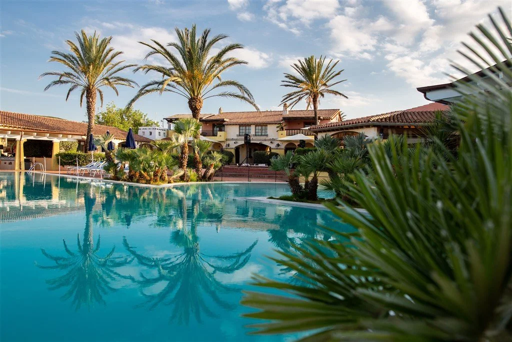 Luxury resort with a large swimming pool, surrounded by palm trees and tropical plants, with buildings featuring terracotta roofs in the background, under a partly cloudy sky.
