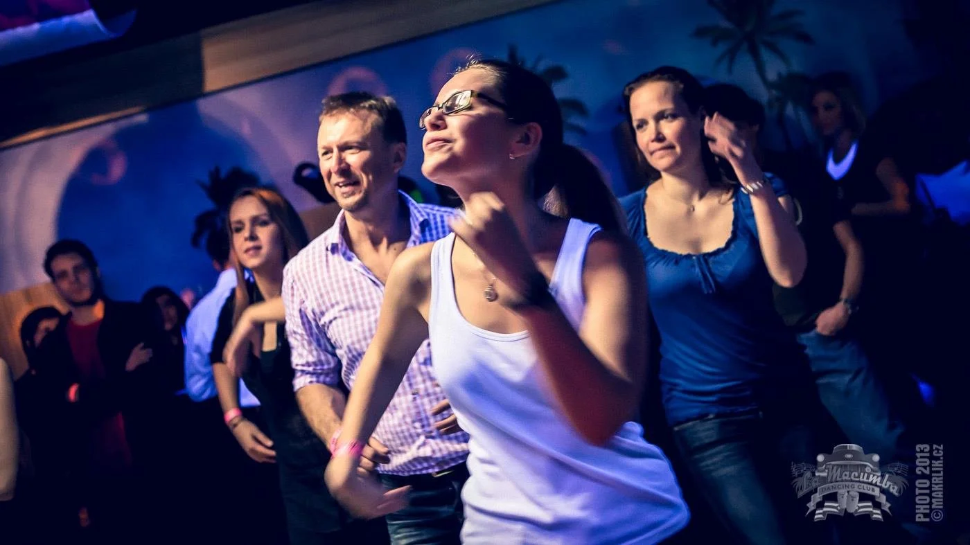Group of people dancing at a nightclub or dance club. The woman in front is wearing glasses and a white tank top, while others are watching or dancing behind her.
