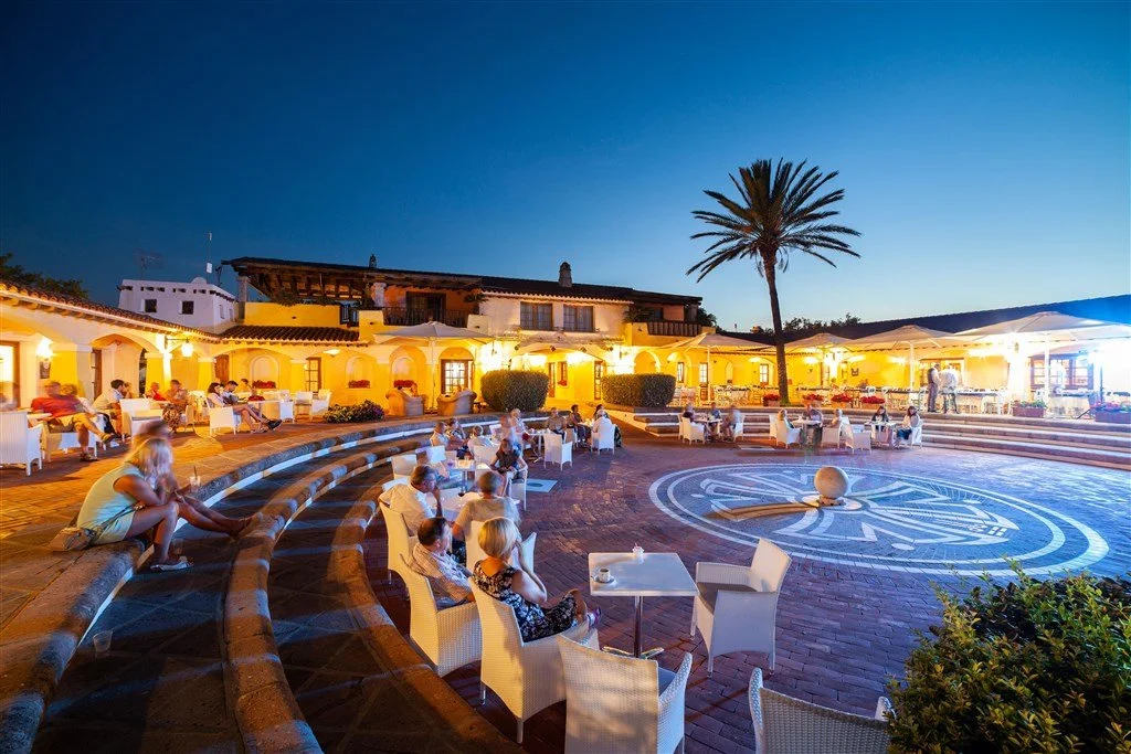 People sitting and relaxing at an outdoor courtyard with a patterned circular design on the ground, illuminated building walls, a large palm tree, and a clear evening sky.