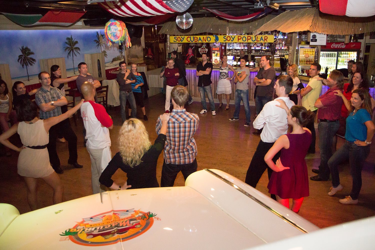 People gathered in a circle on a wooden floor, socializing and dancing in a bar with colorful decorations, tropical mural, and a sign that reads "Soy Cubano Soy Popular", with a bar counter in the background.