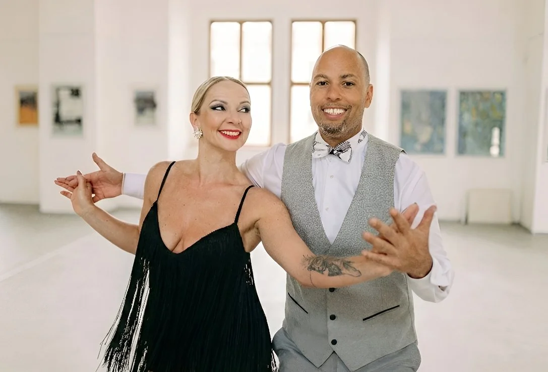 A man and woman dancing together in a brightly lit, spacious room with large windows and artwork on the walls.