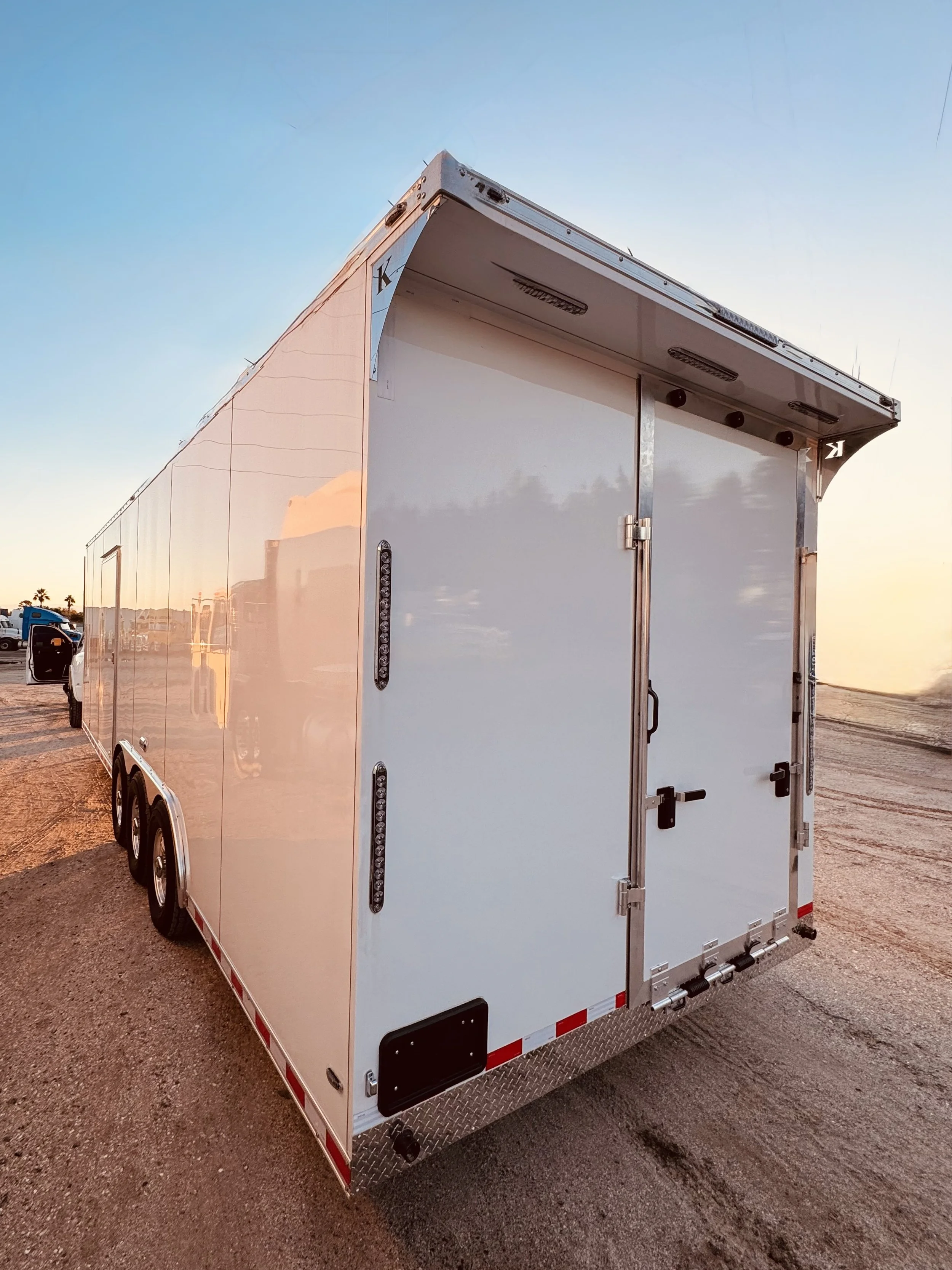 White enclosed trailer with double rear doors, set on a dirt lot at sunset, reflecting the sky and surroundings.