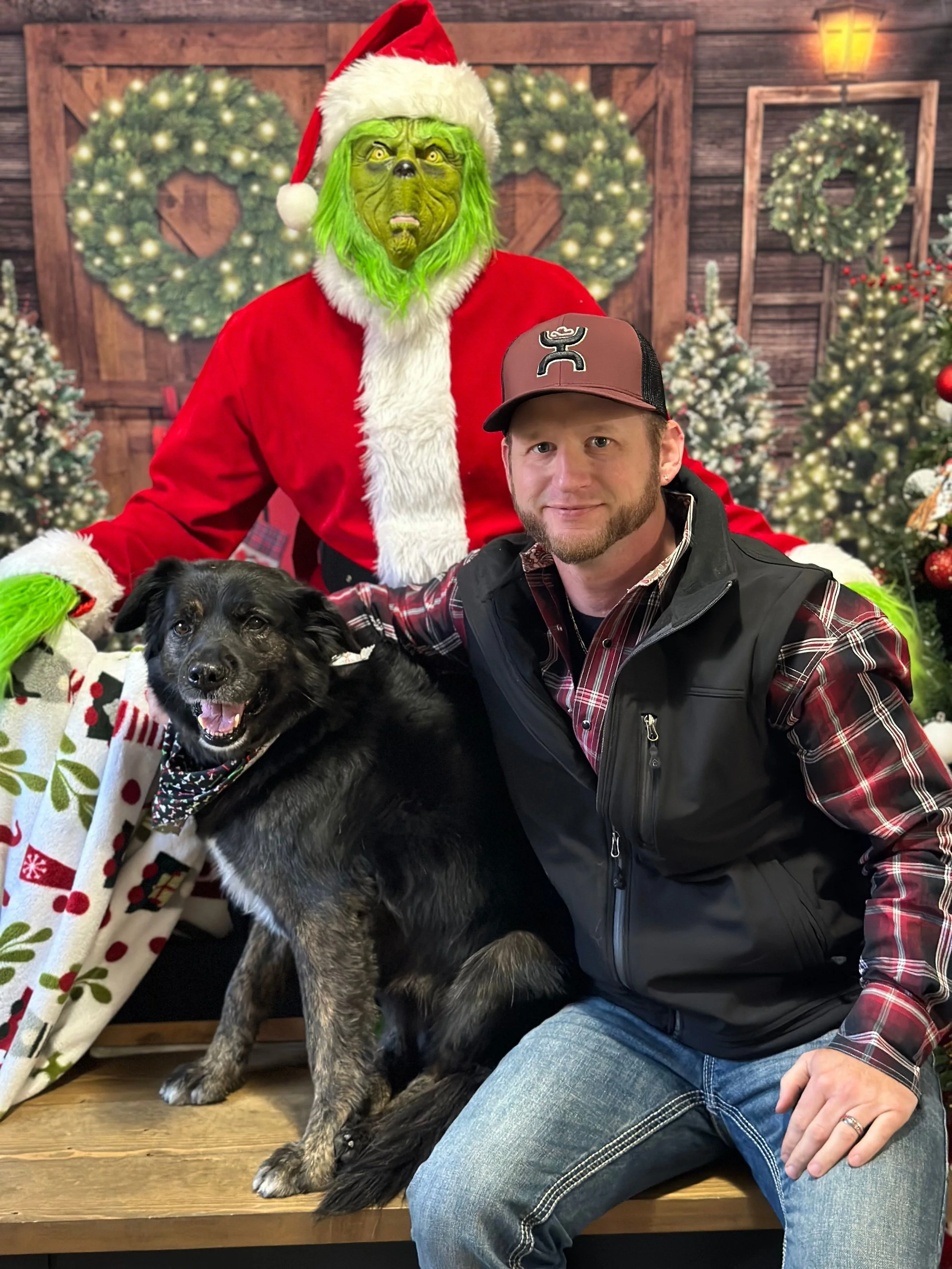 A man, a dog, and a green-faced creature in a Christmas scene with decorated trees and wreaths. The man is wearing a plaid shirt, vest, and a baseball cap, and is sitting on a bench. The dog is sitting beside him, wearing a festive bandana, and the creature dressed as the Grinch in a Santa costume with a hat has a green face and hands.