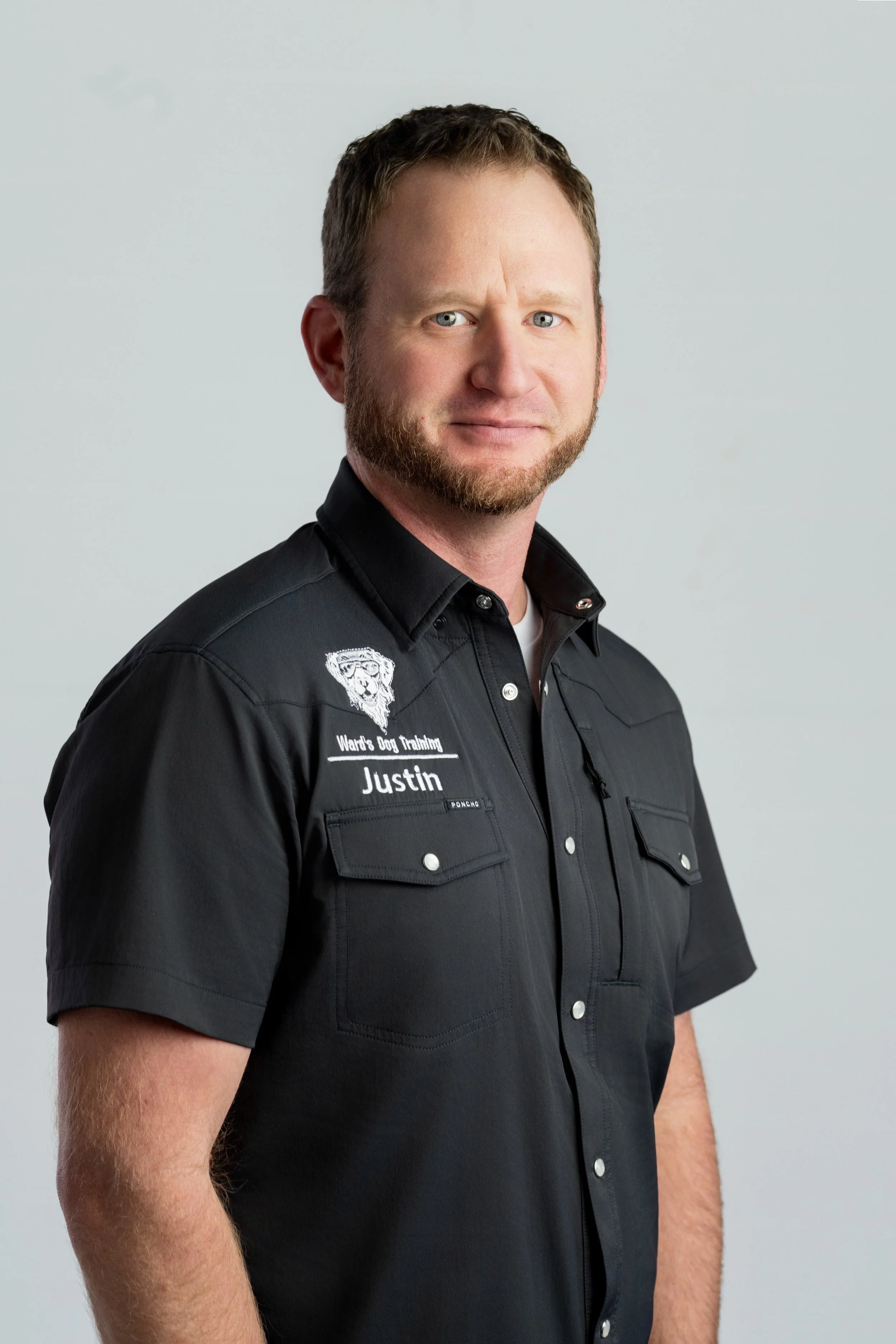 Portrait of a man, Justin Ward, the owner  in a black shirt with patches, standing against a plain background.