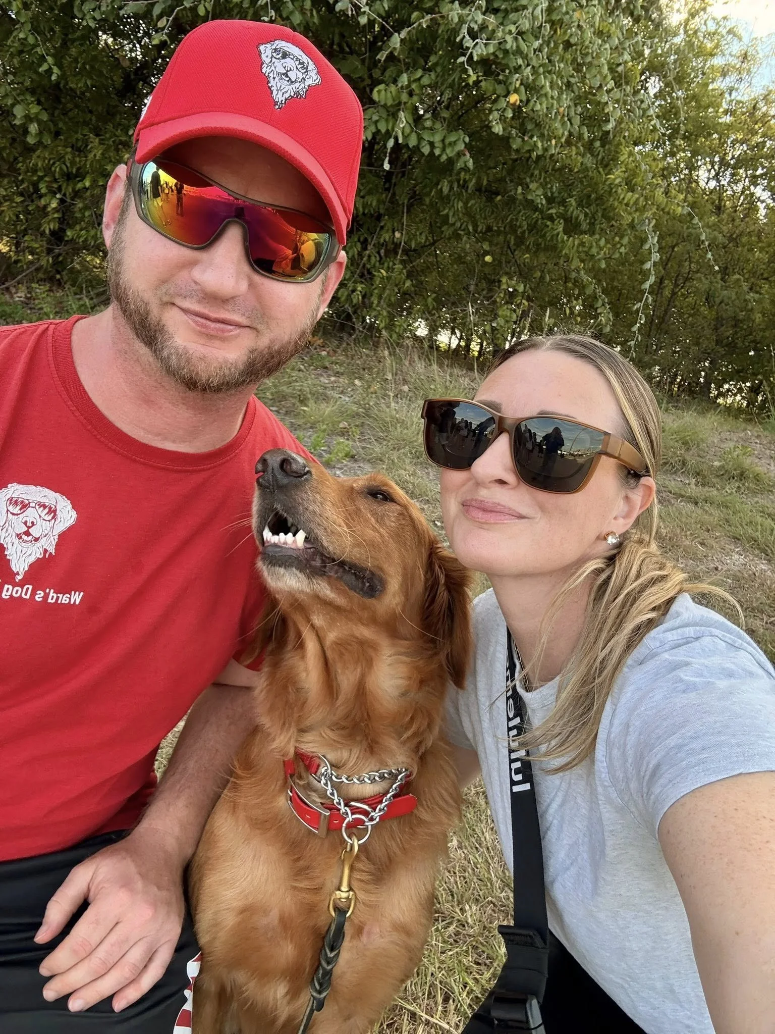 A man and woman with a dog outdoors during daytime, all wearing sunglasses. The man is wearing a red cap with a logo, and a red T-shirt with a matching logo. The woman has blonde hair tied back and is wearing a gray T-shirt. The dog is a golden retriever with a red collar and chain and appears to be mid-bark or squeak with its mouth open.