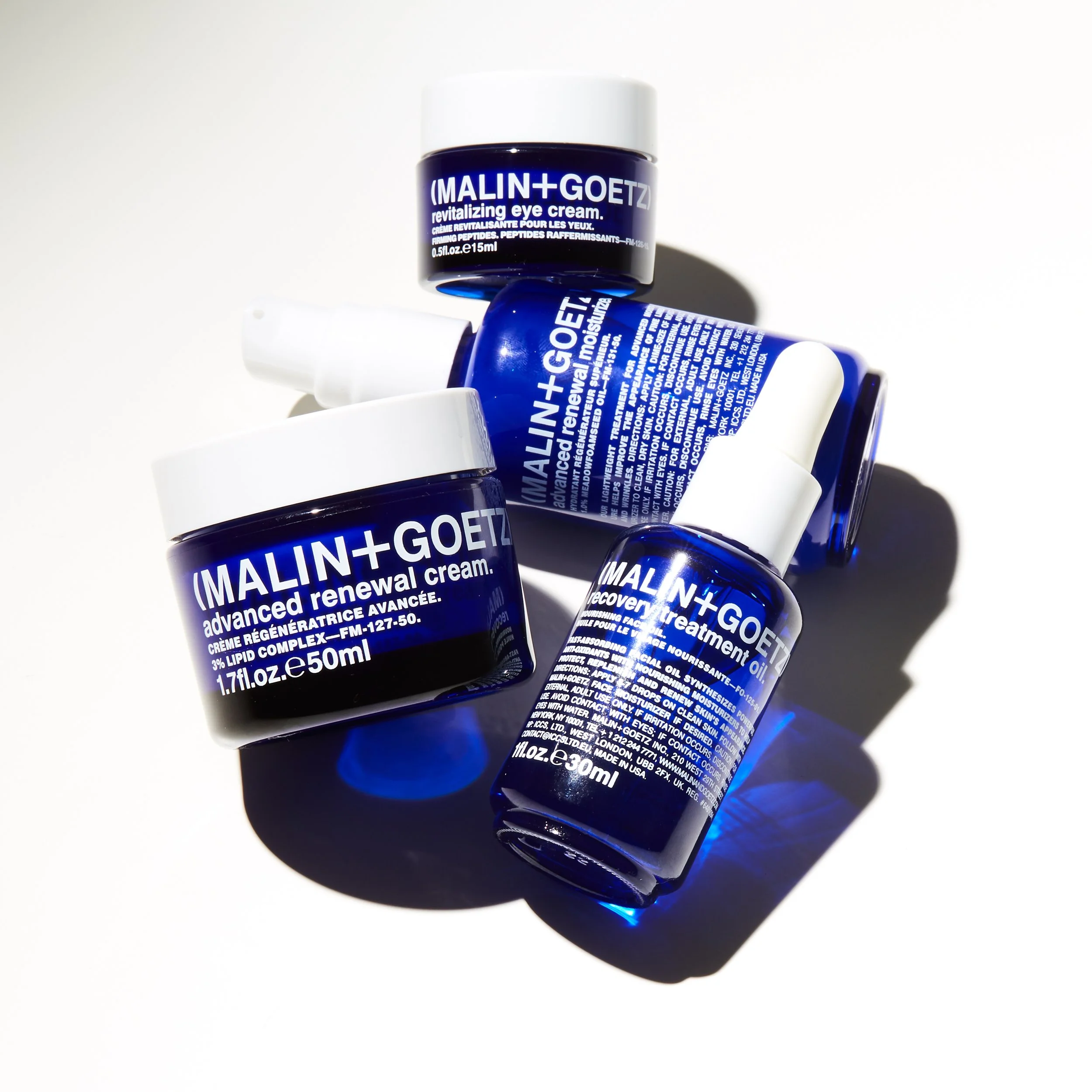 Variedad de frascos de productos Malin+Goetz para cuidado facial, incluyendo crema revitalizante para los ojos, crema de renovación avanzada y tratamiento facial, en envases azul con etiquetas blancas sobre fondo blanco.