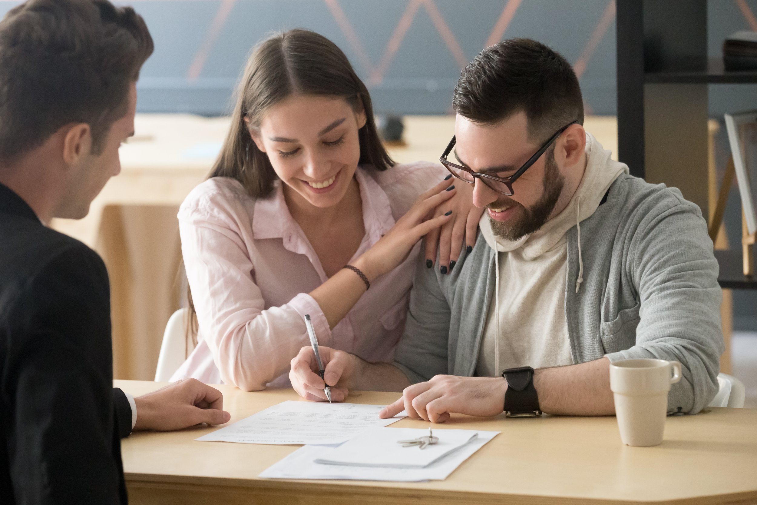 Three young adults sitting at a table, smiling and signing a document with a pen, during a casual meeting or signing ceremony, with a coffee mug on the table.
