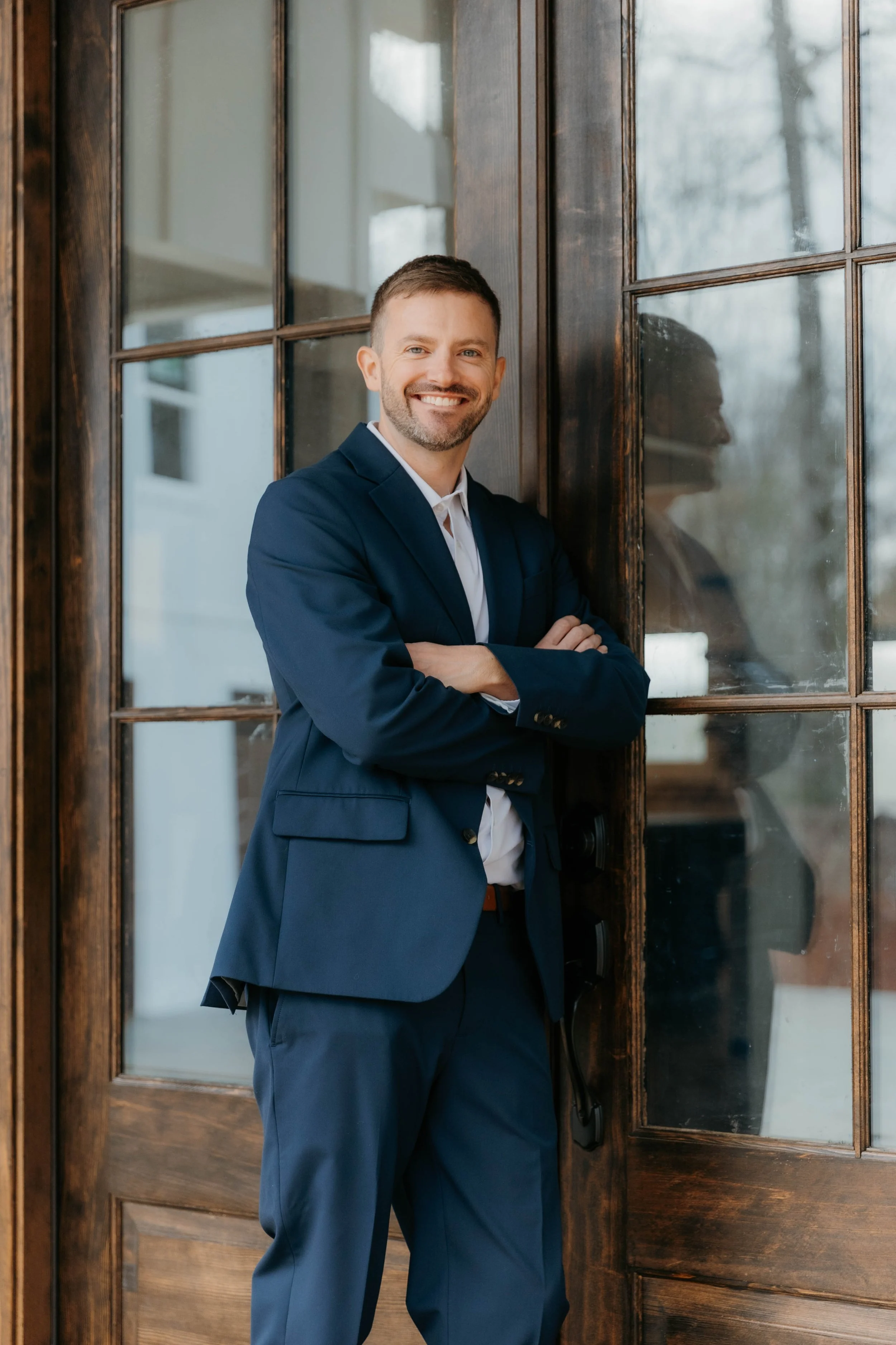 Man in a navy suit leaning against a wooden door with glass panels, smiling with arms crossed.