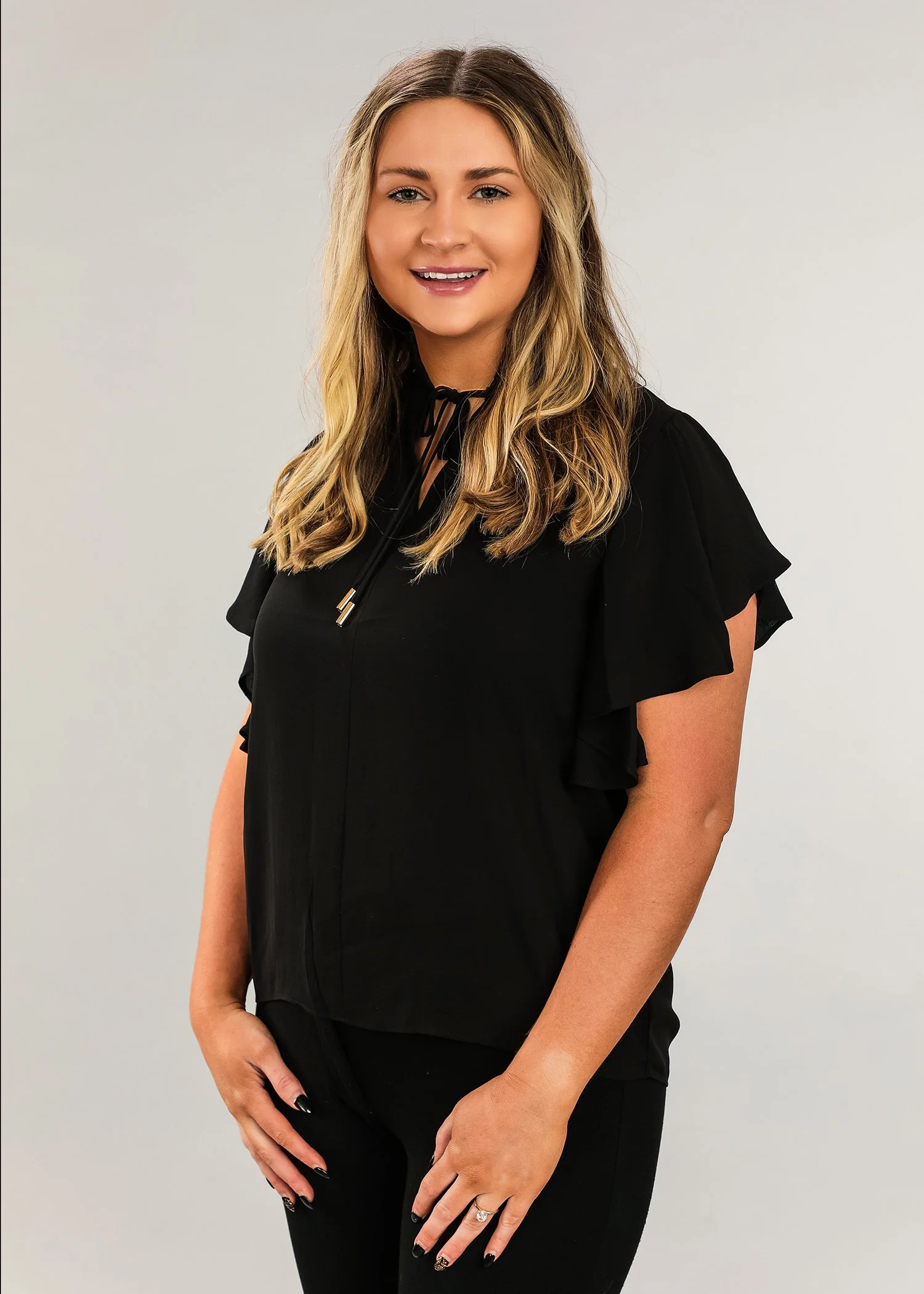 A woman with blonde hair wearing a black top and black pants, smiling and standing against a light gray background.