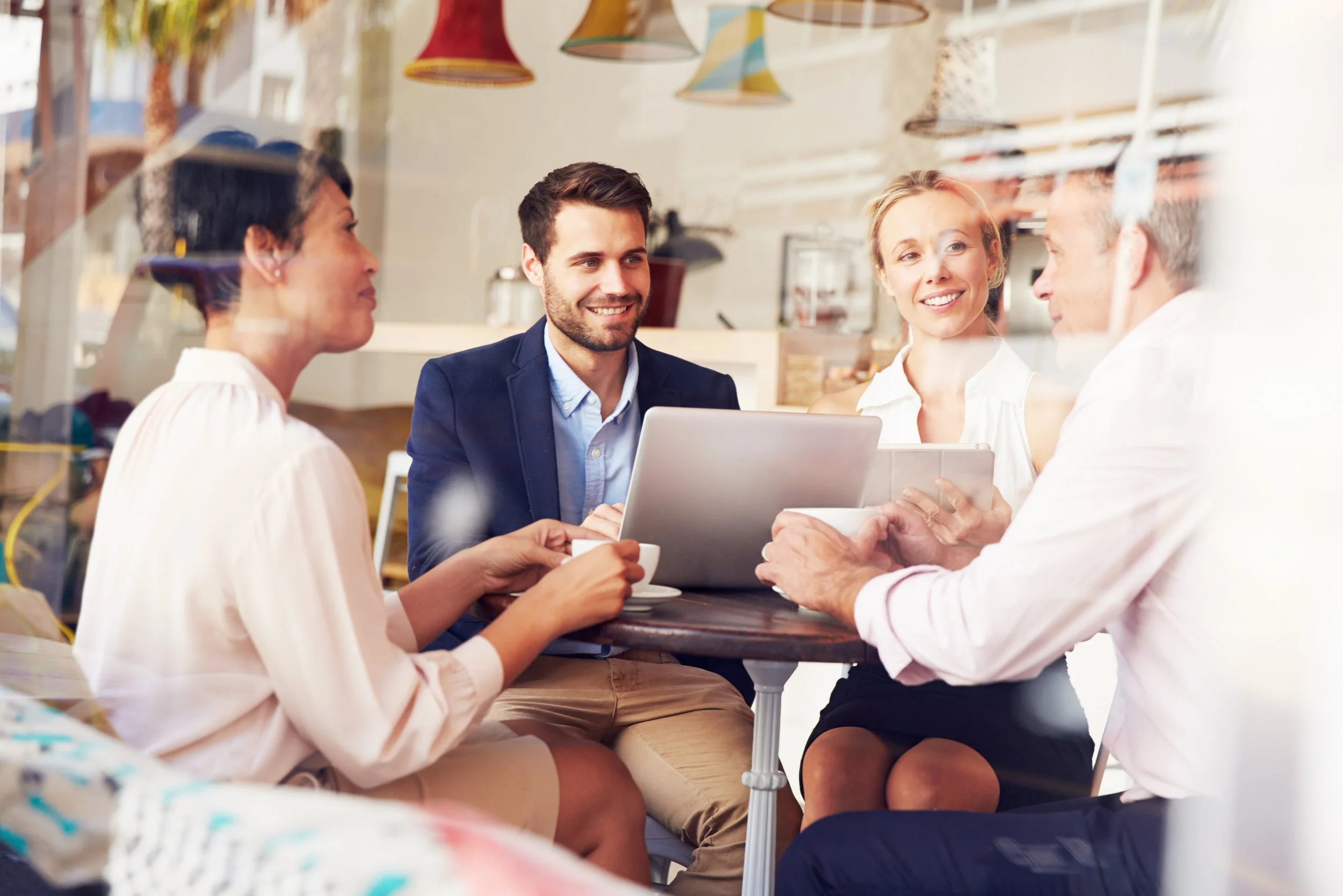 Four people sitting around a table in a cafe, engaged in conversation and looking happy. Two women and two men, with one woman holding a cup and the others with a laptop and tablet.