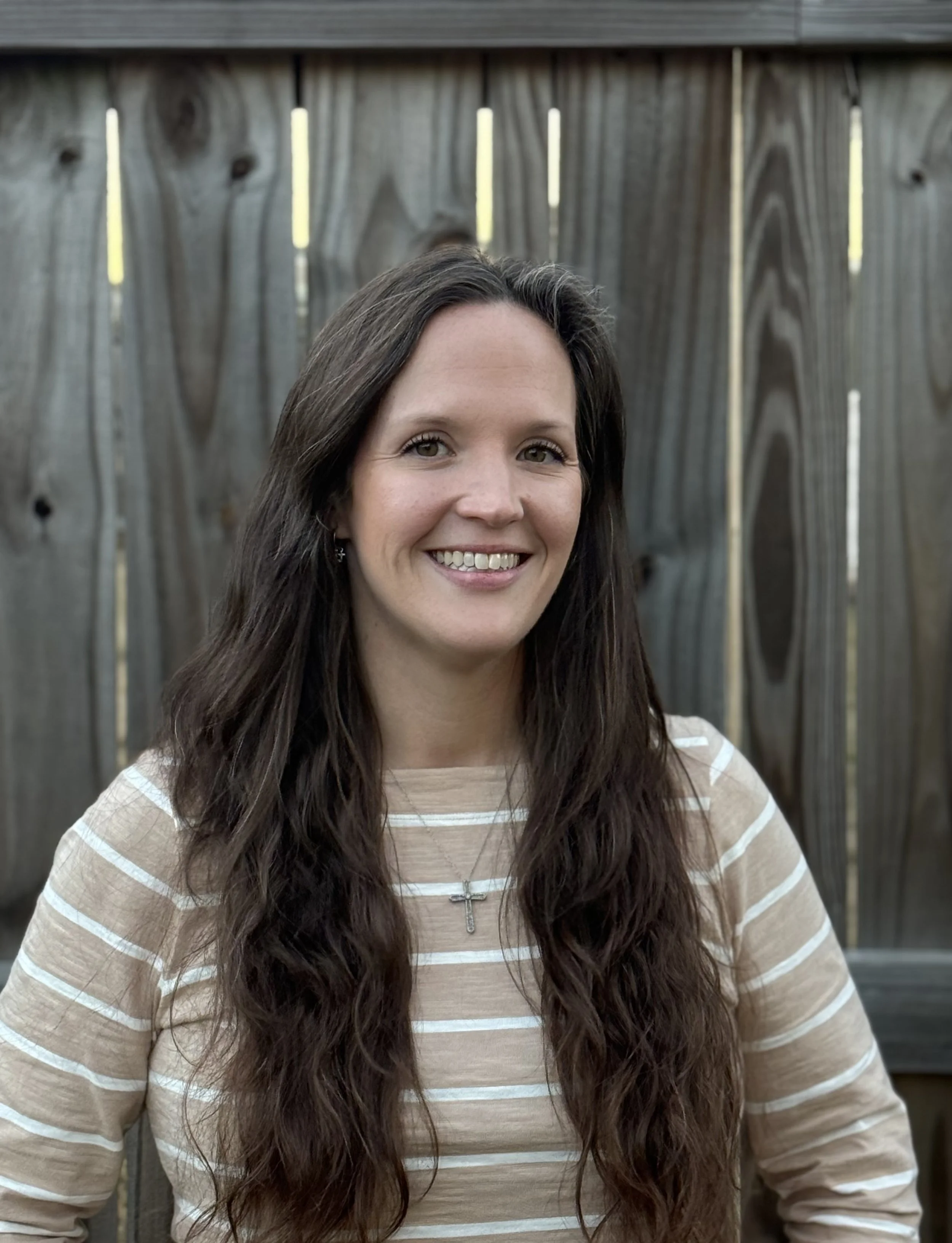 A woman with long brown hair smiling outdoors in front of a wooden fence, wearing a beige and white striped shirt and a cross necklace.