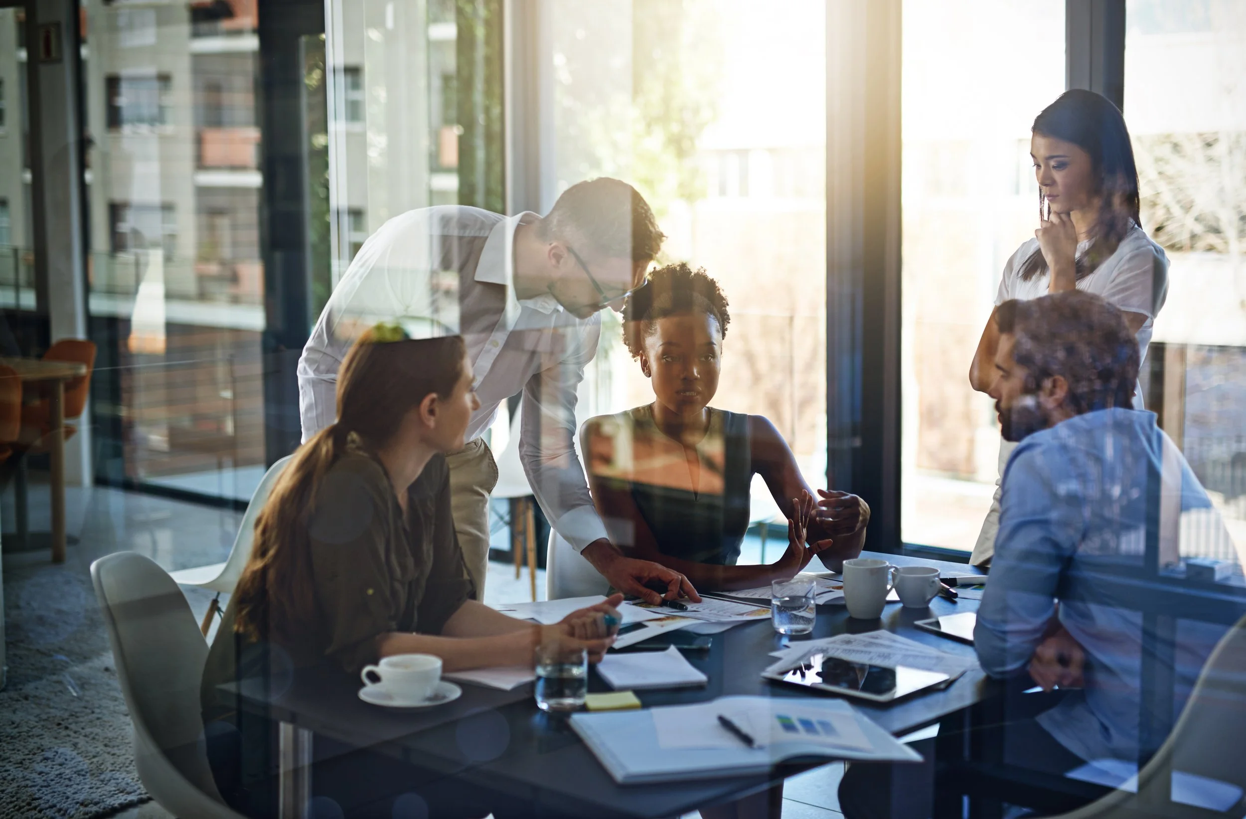 Team of five professionals in a meeting room with large windows, discussing documents and working on a project.