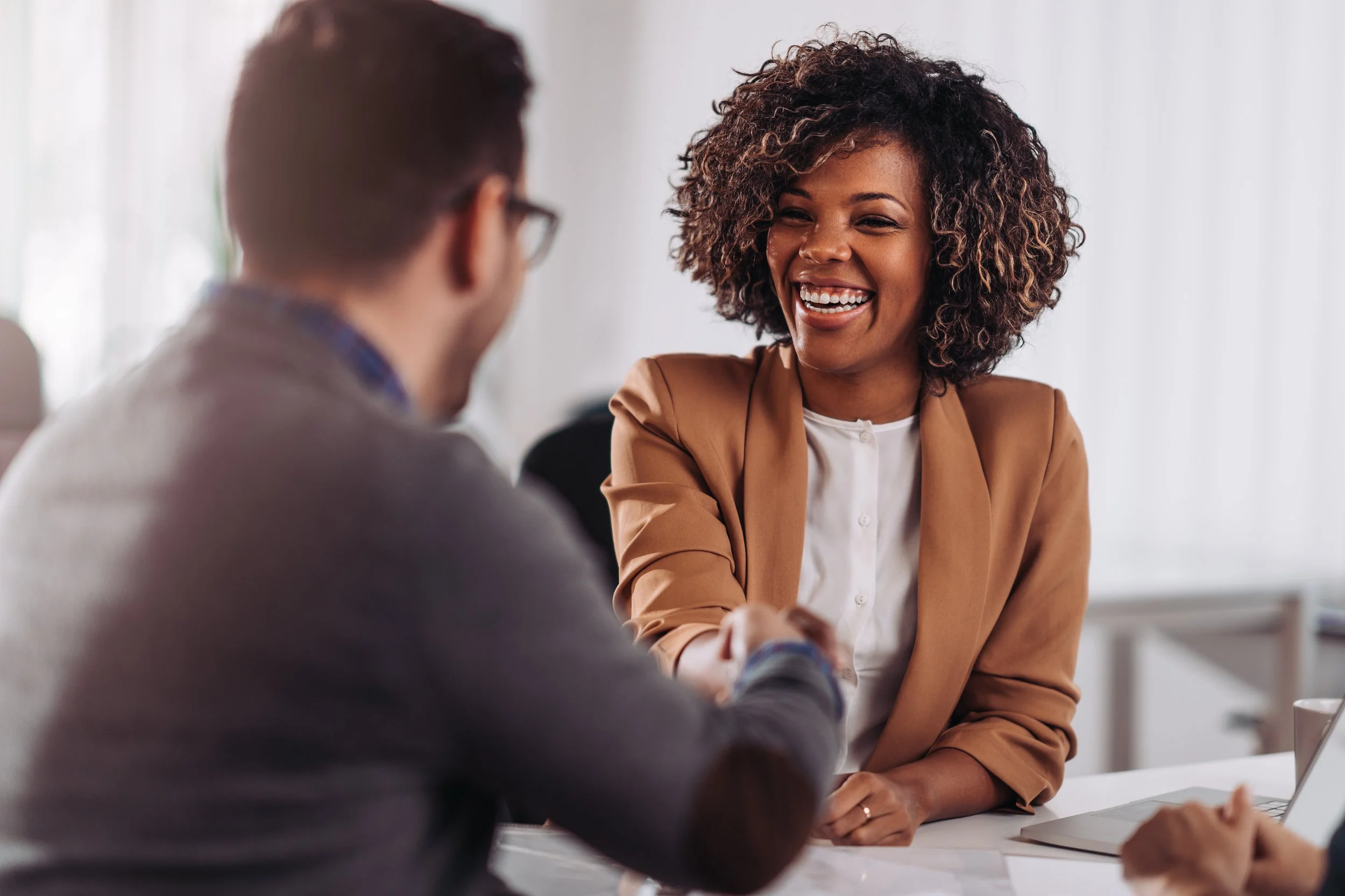 A woman with curly hair smiling and shaking hands with a man in glasses, in a bright office setting.