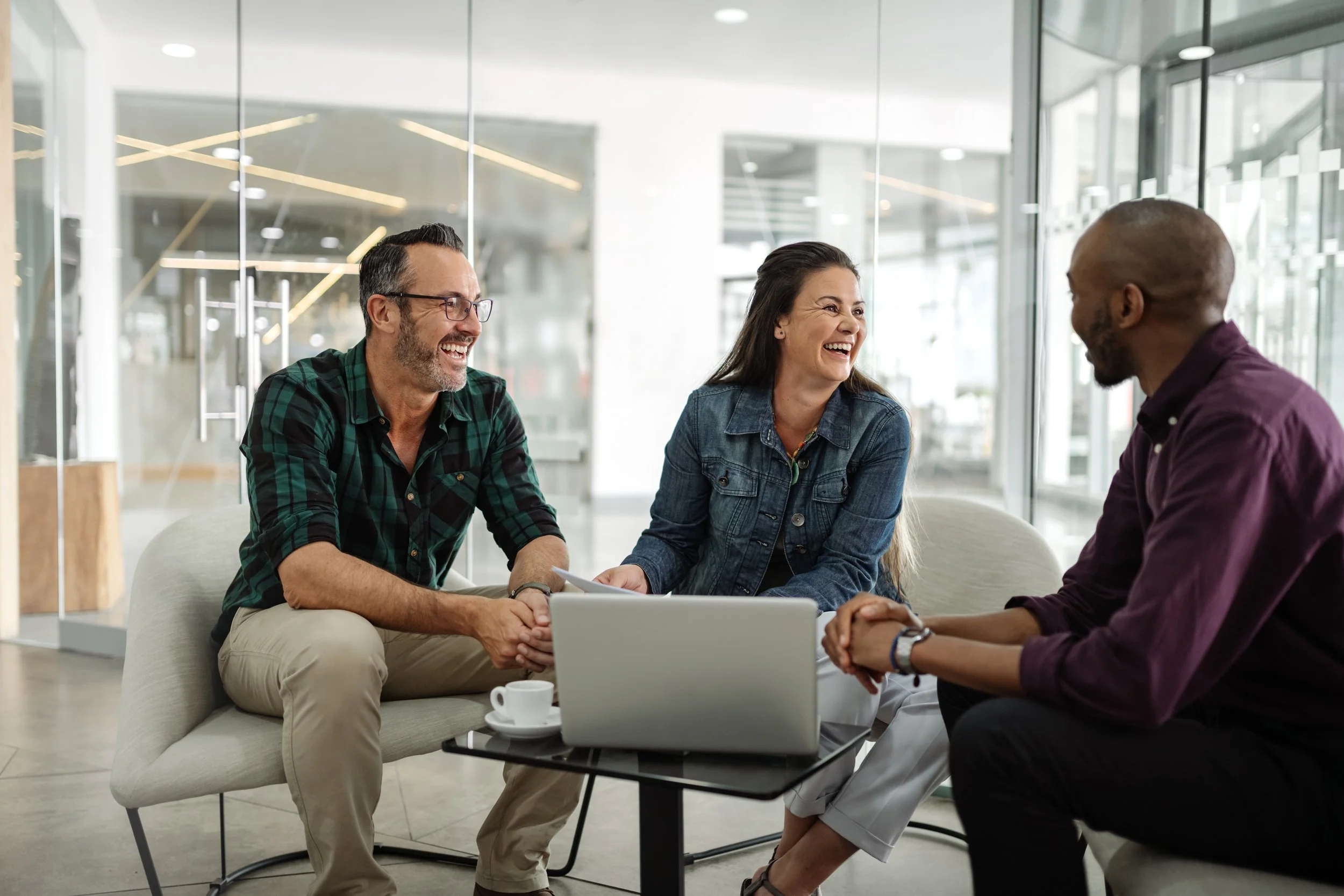 Three people sitting on a couch, smiling and talking, in a modern office lounge with glass walls and a laptop on a table in front of them.