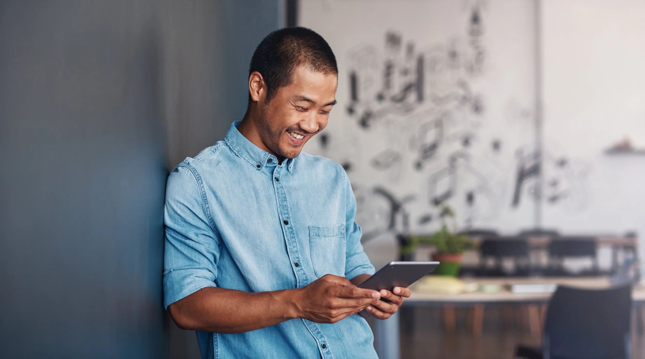A man is smiling while looking at his smartphone, standing in a modern office space with a wall art background and a conference table in the distance.