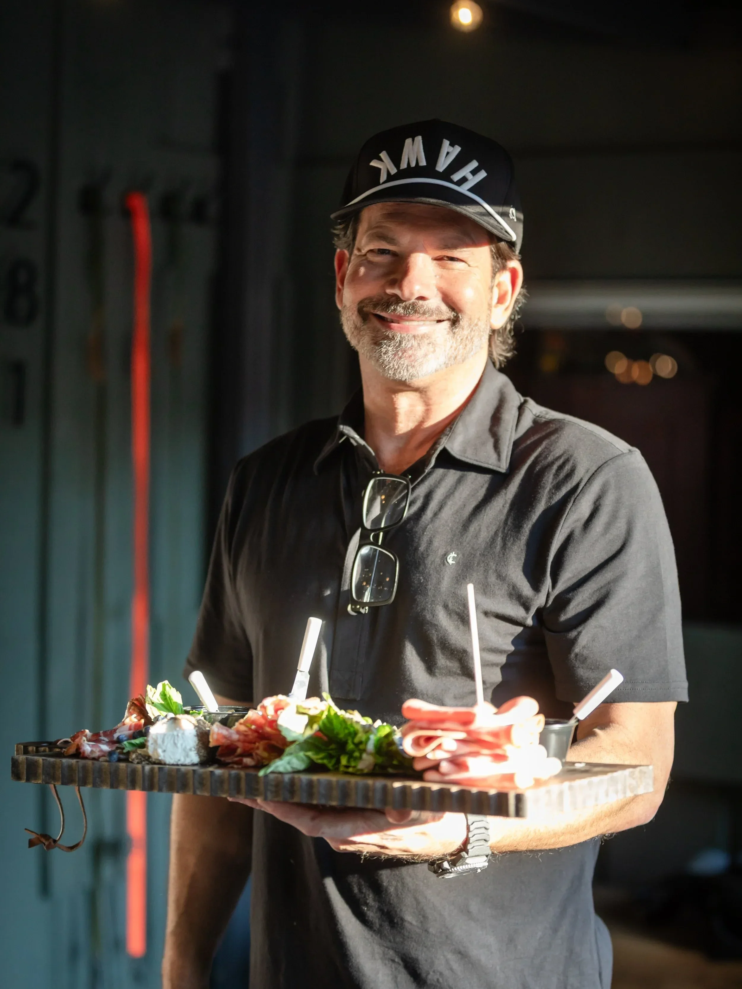 A smiling man with glasses hanging from his shirt collar holding a tray of assorted food, including meats, cheese, greens, and small cups with straws, in a dimly lit setting.