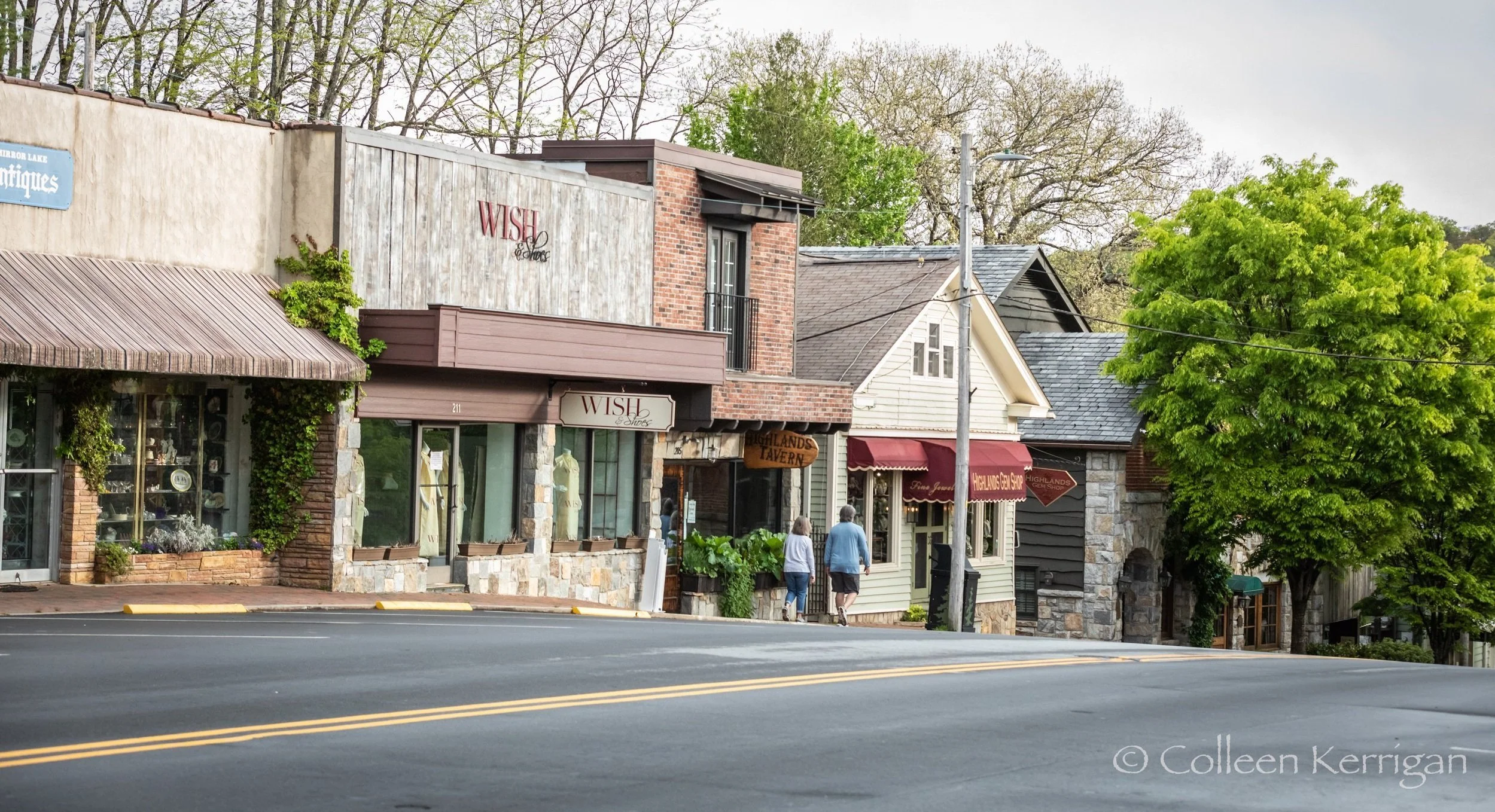 A small town street view with buildings, trees, and two people walking on the sidewalk.