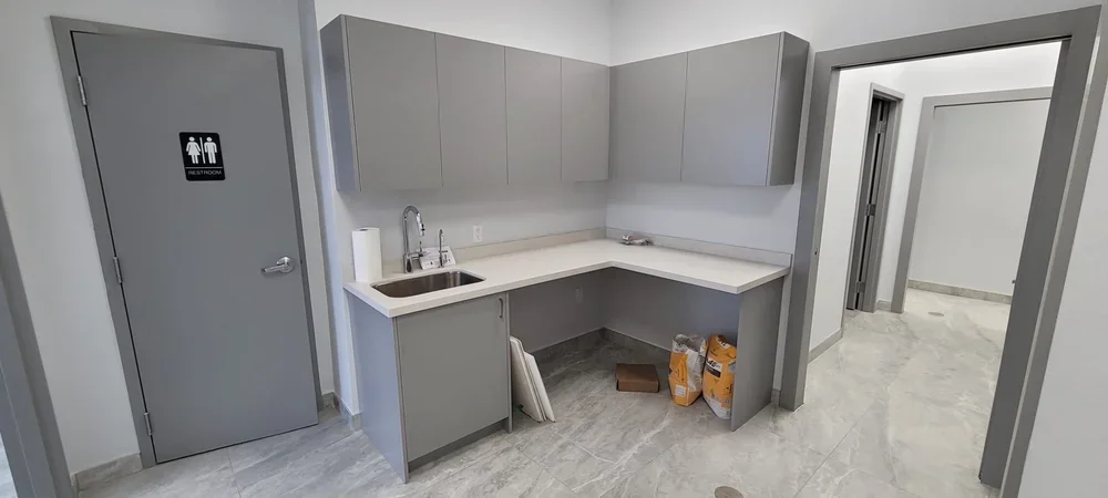 Grey door with restroom sign, grey cabinets, white countertop with sink, paper towel roll, and construction materials on the floor in a small kitchen or utility room.