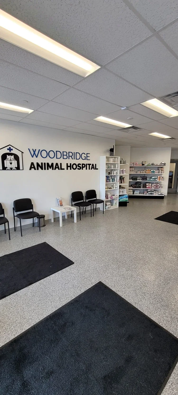 Waiting area at Woodbridge Animal Hospital with black chairs, a small white table, shelves with pet supplies, and the hospital's sign on the wall.