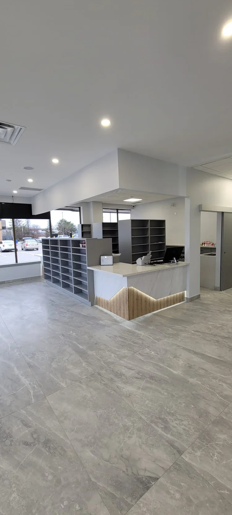 Empty reception area with gray shelves, a counter with a computer and payment terminal, large windows in the background, and gray marble flooring.