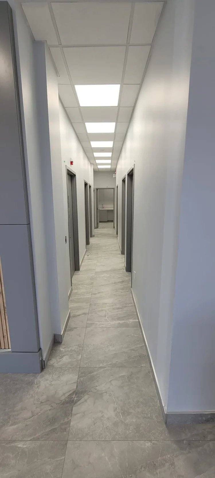 Empty hospital corridor with four doors, gray walls, tiled gray flooring, and ceiling lights.