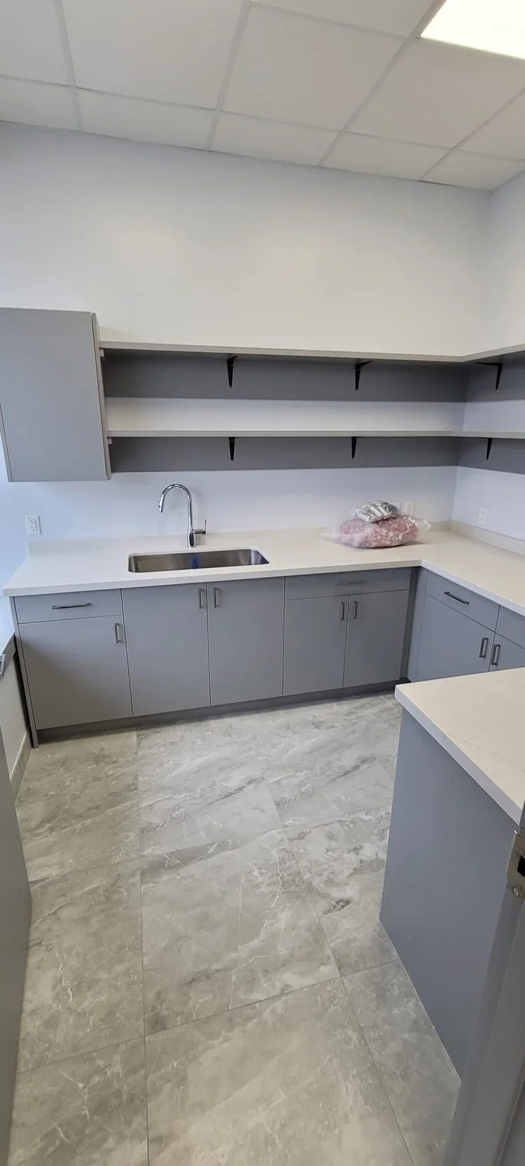 Modern kitchen with gray cabinets, beige countertops, a stainless steel sink with a faucet, open shelves above, and a gray tiled floor.