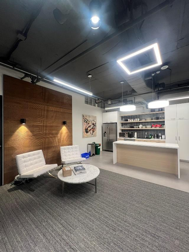 Modern office lounge area with white chairs, a marble coffee table, artwork on the wall, and an open kitchen with shelves and appliances.
