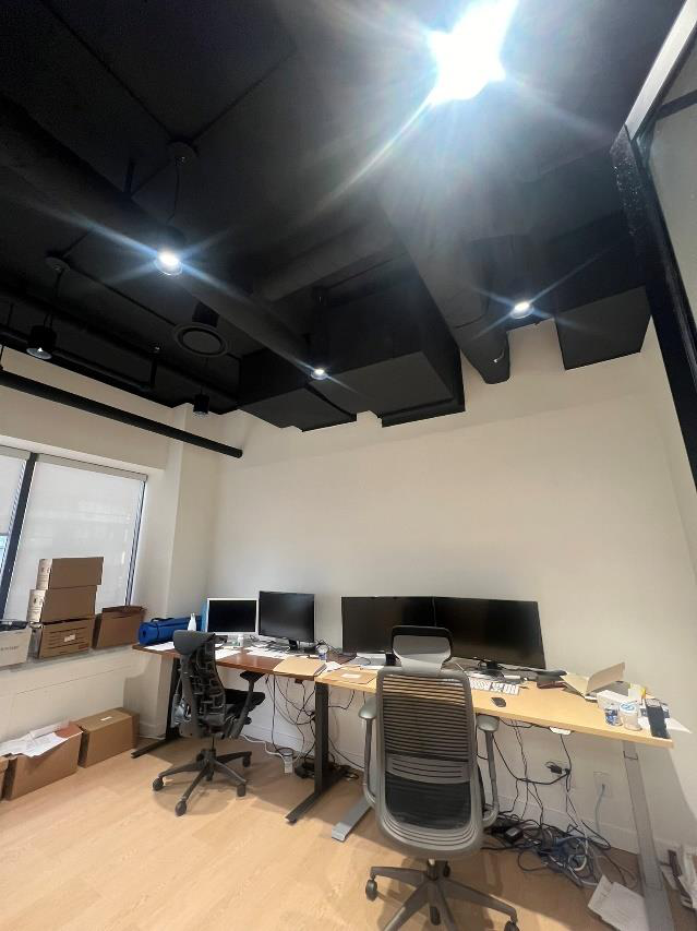 An office with three computer monitors on a long desk, two office chairs, and cardboard boxes on the window sill. Overhead ceiling lights are turned on, illuminating the room.