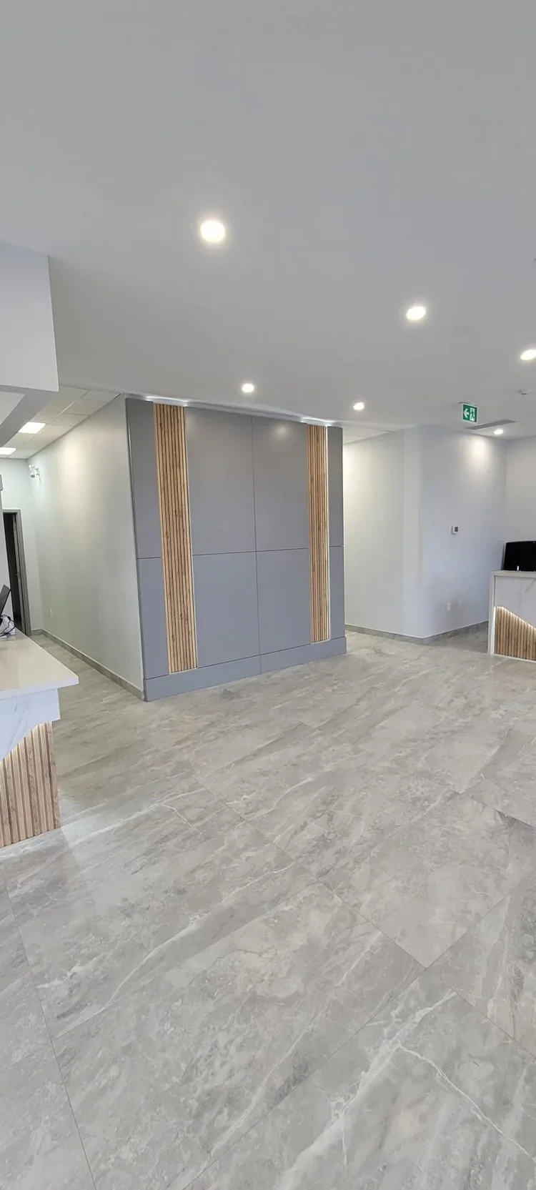 Modern interior space with gray tiled floors, white walls, and ceiling with recessed lighting. There's a gray partition with wooden accents, and a small black computer monitor is visible on the right.