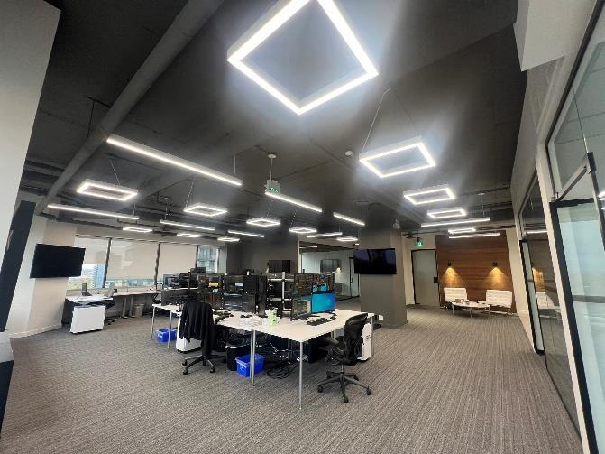 Modern office with workstations, multiple computer monitors, and LED ceiling lights, along with a small seating area in the background.