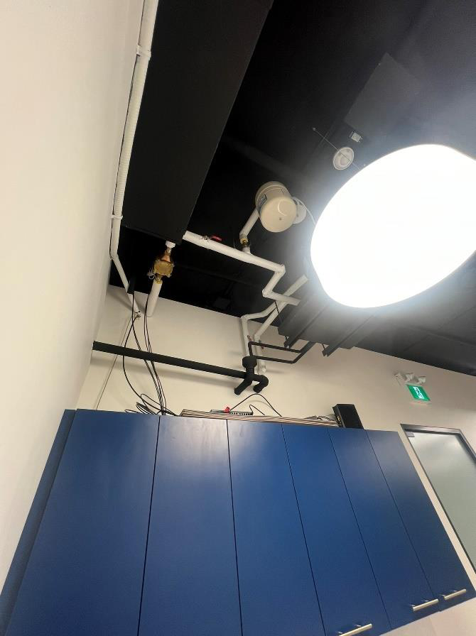 Ceiling with black panels, white pipes, a large bright ceiling light, and a blue storage cabinet below.