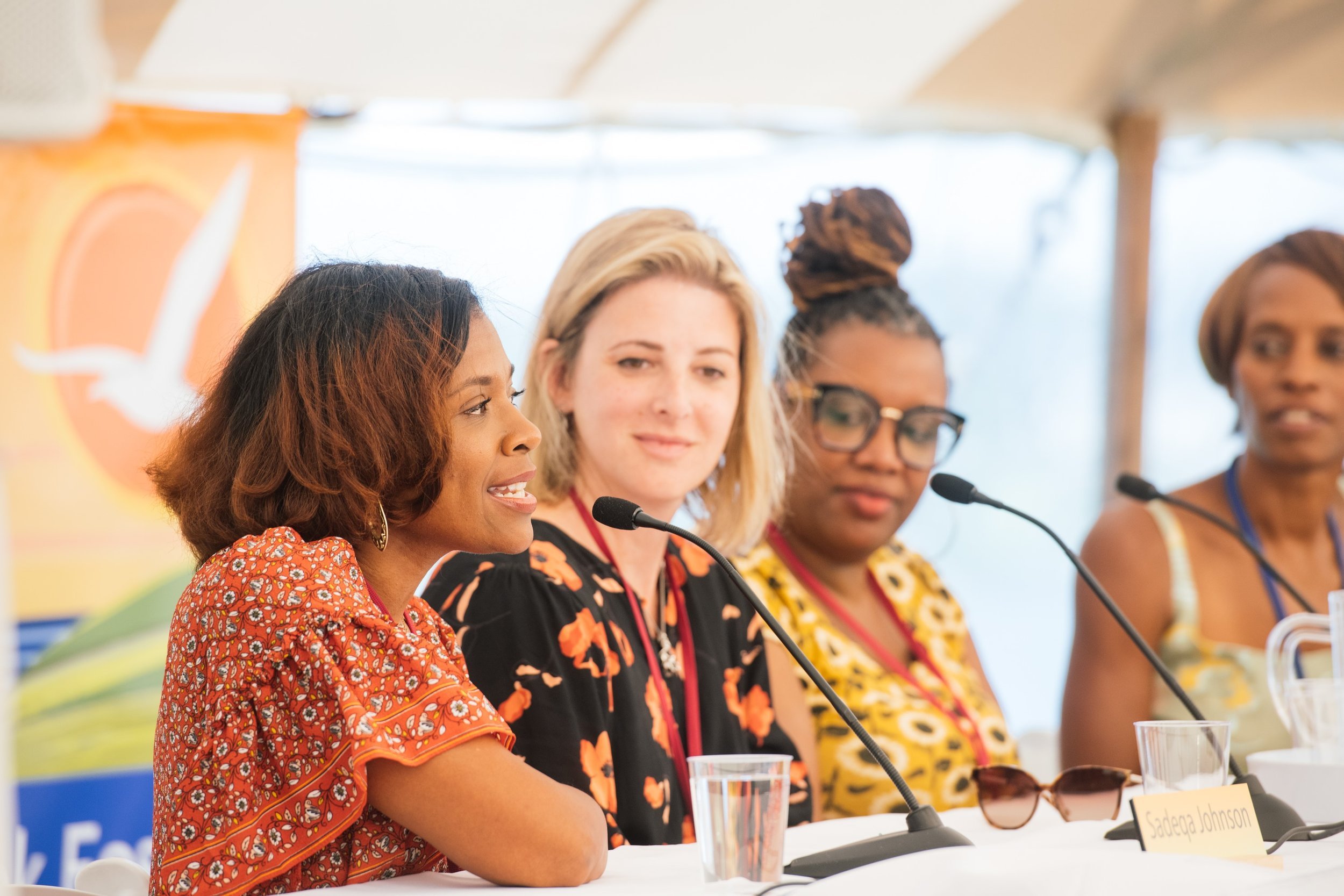 Four women sit at a panel discussion, each with a microphone in front of them. The woman on the left is speaking, wearing a red patterned top. The other women are listening; one with blonde hair, another with glasses and a bun, and the last with short hair.