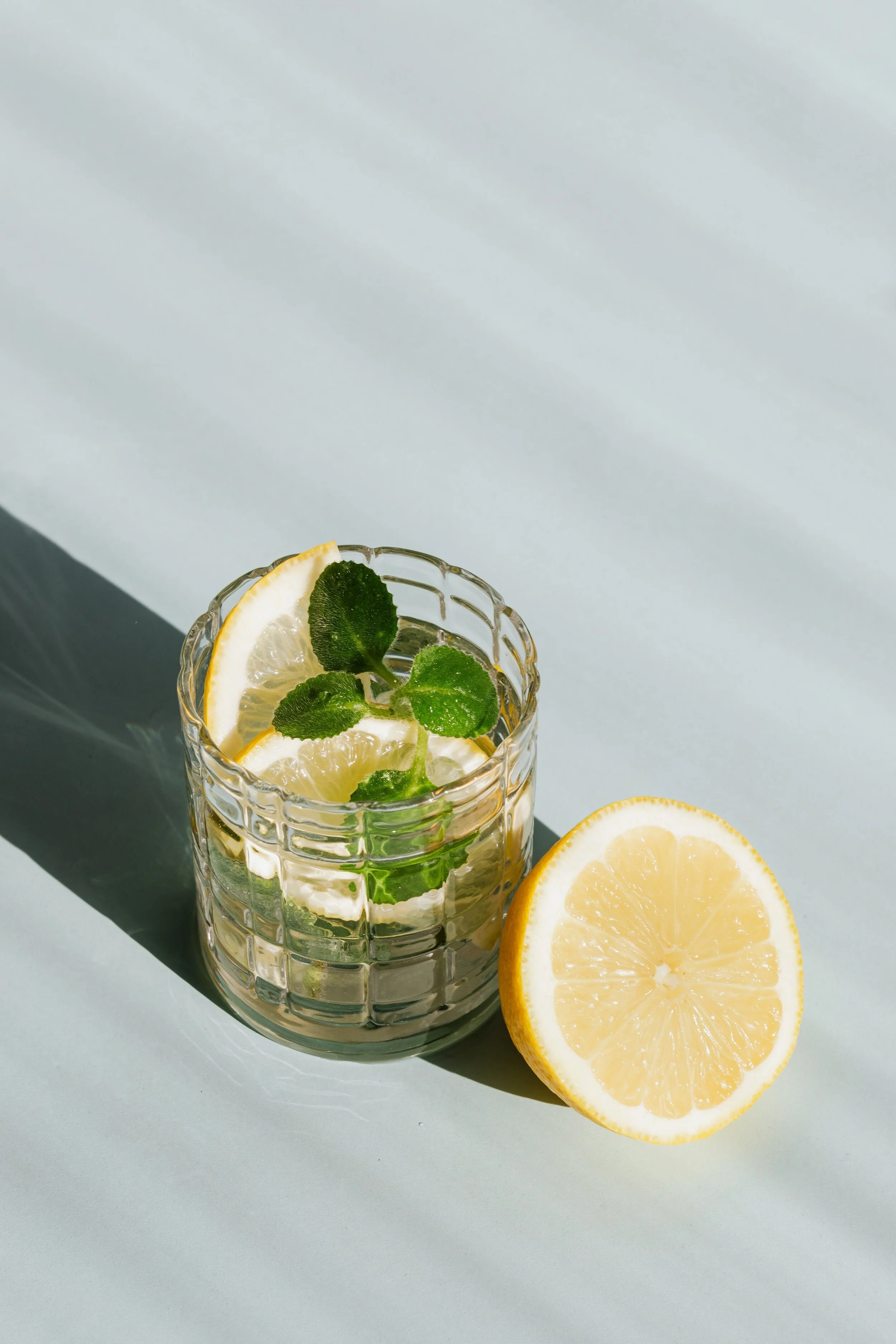 A glass of lemonade with lemon slices and mint leaves next to a halved lemon on a light green surface