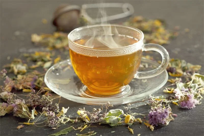 A steaming cup of herbal tea in a clear glass cup on a matching saucer, surrounded by dried flower petals and herbs on a dark surface.