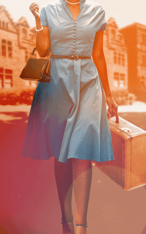 A woman in a blue dress holding a vintage suitcase and a small black purse, standing outdoors with buildings in the background.