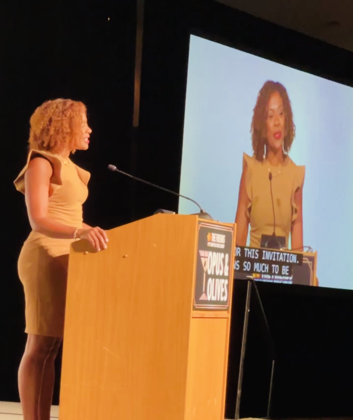 A woman with curly blonde hair standing behind a wooden podium on stage, presenting at an event. She is wearing a tan dress and speaking into a microphone, with a large screen displaying her image in the background.