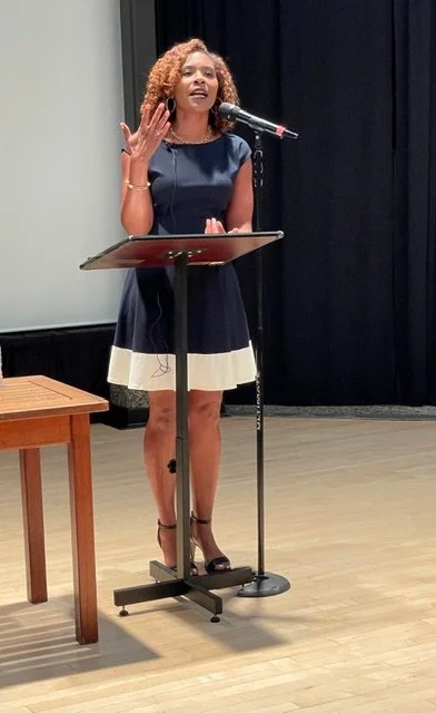 A woman with curly hair speaking at a podium with a microphone in a room with a black curtain background.