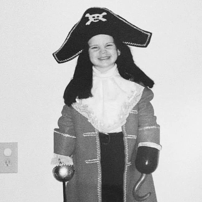 Justin Girard as a child dressed as a pirate with a pirate hat featuring a skull and crossbones, smiling with a toy sword and hook, wearing a pirate costume with a coat and accessories.