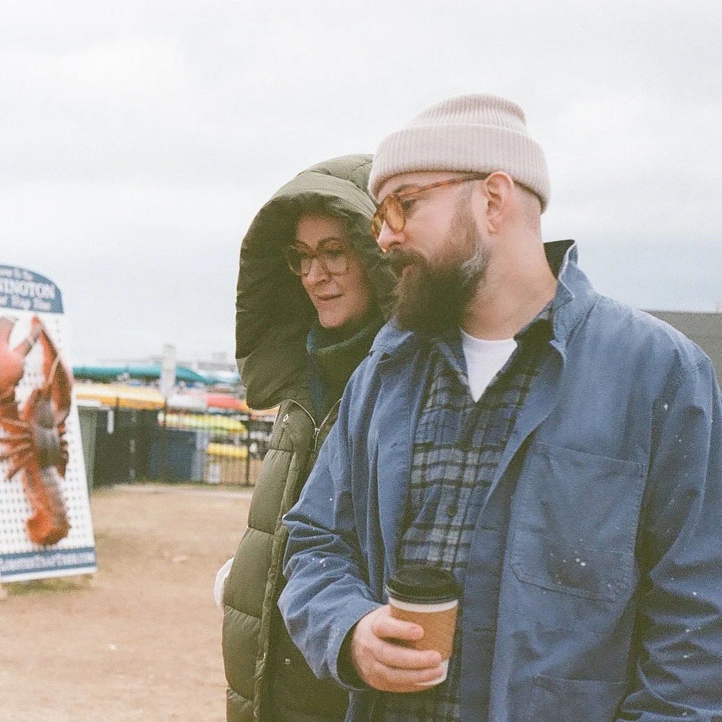 Justin Girard, wearing glasses, a white beanie, and a blue jacket, holding a coffee cup, standing outdoors with a woman in a hooded coat behind him at a fair or outdoor market.