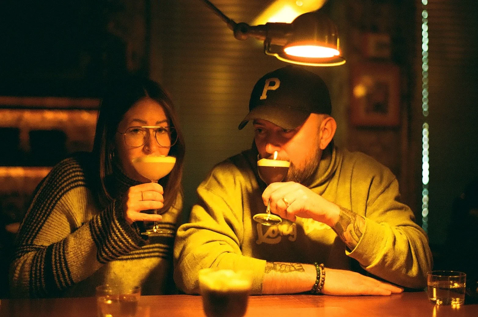 Justin Girard sitting at a bar, with his wife, drinking cocktails under dim, warm lighting. The woman on the left has glasses and dark hair, and the man on the right is wearing a baseball cap and has tattoos.