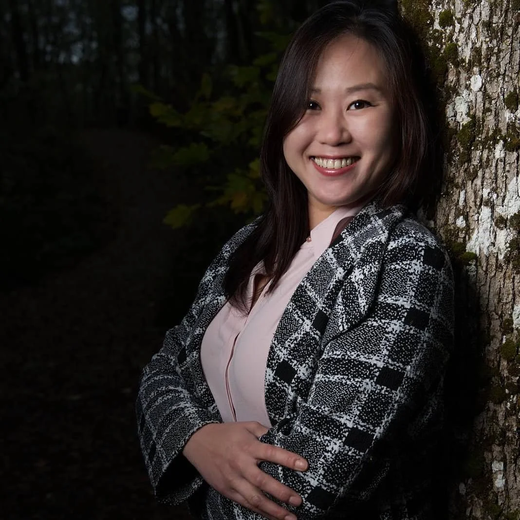 A woman with long brown hair smiling and leaning against a tree in a forest, wearing a checkered blazer and pink blouse.