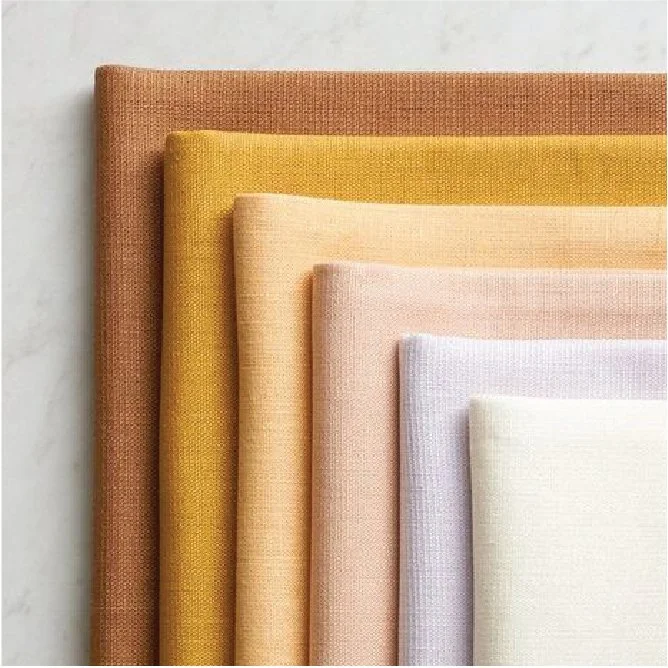 Arrangement of seven folded textured fabric pieces in shades of brown, yellow, peach, lavender, and white, layered on top of each other on a light marble surface.