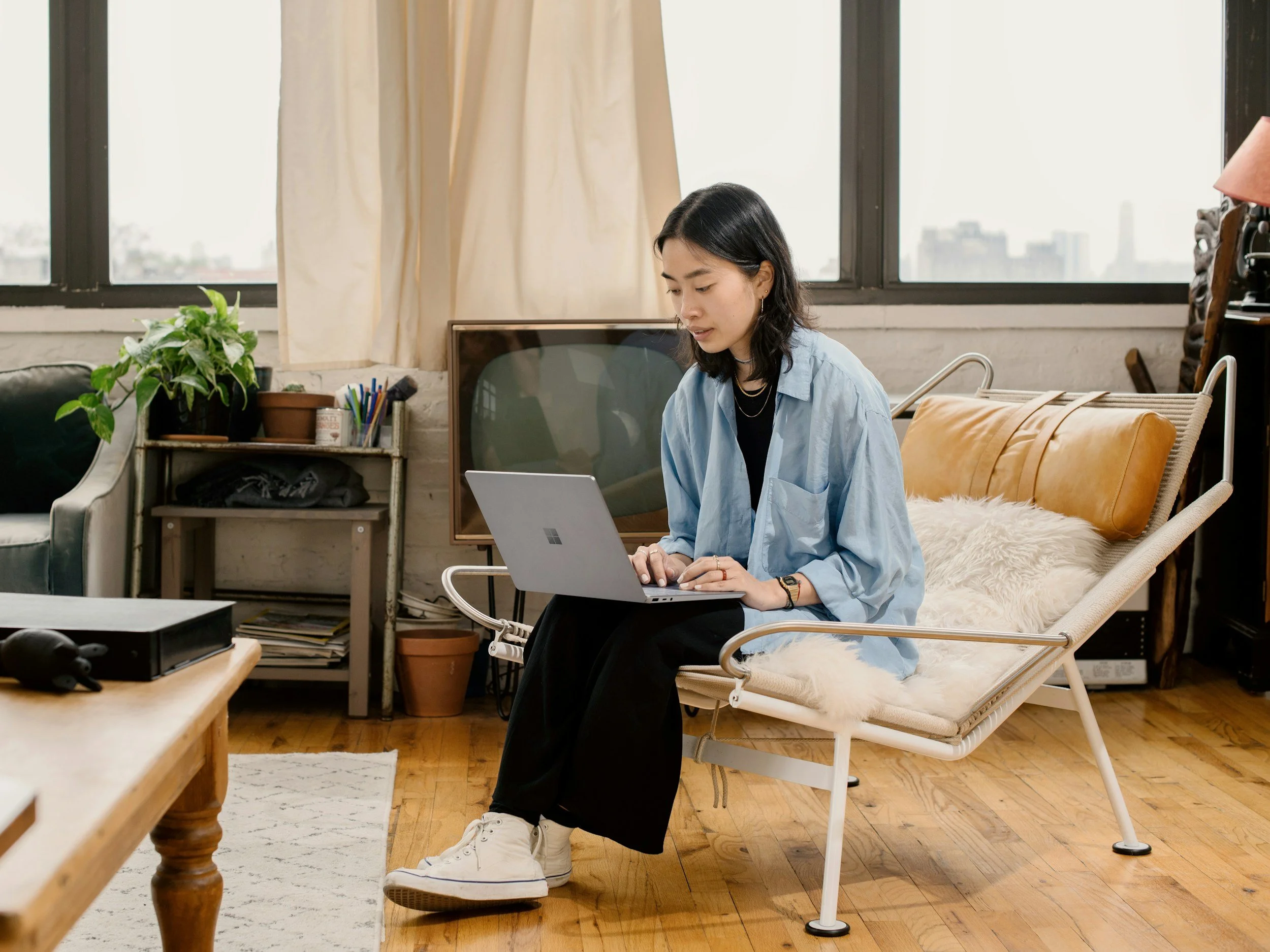 A woman working on a silver laptop while sitting on a white lounge chair with a fur cushion in a cozy apartment with large windows and city view.