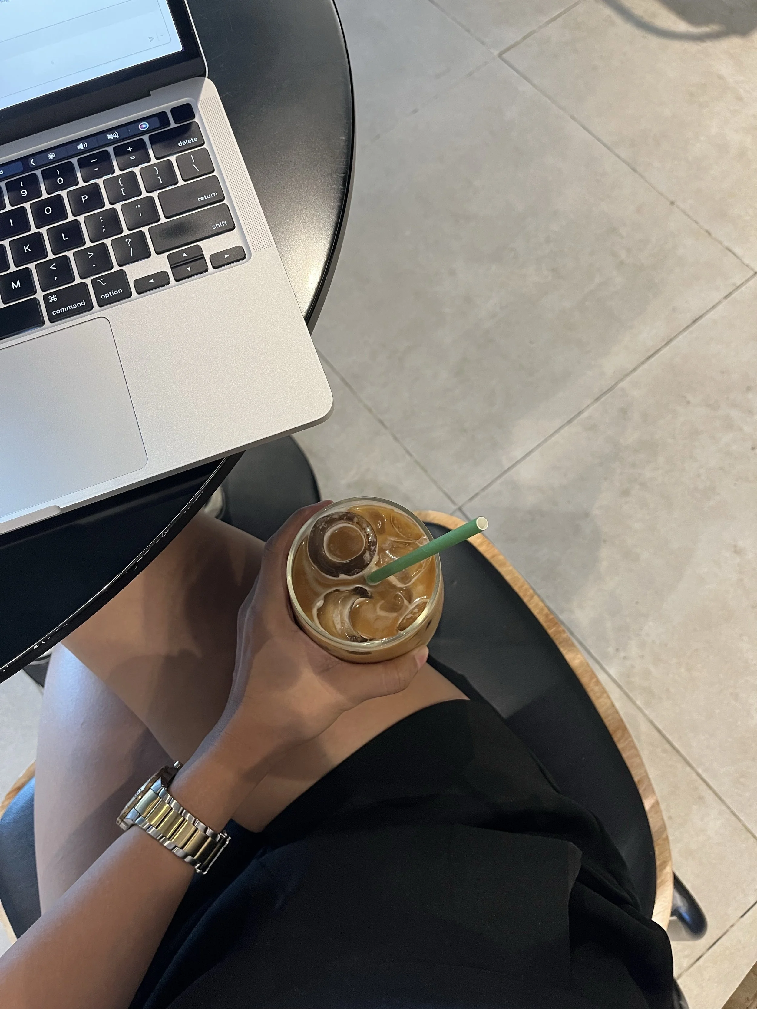 Person sitting at a table with a silver laptop and holding a clear plastic cup of iced coffee with ice and a green straw. The person is wearing a silver watch and black clothing, with a tiled floor visible in the background.