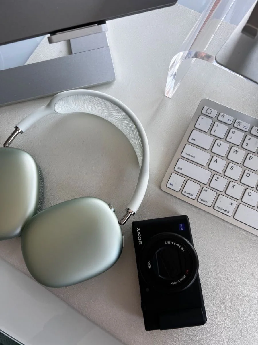 Close-up of a white desk with a computer monitor, a pair of green headphones, a Sony digital camera, a white keyboard, a glass of water, and a computer screen.