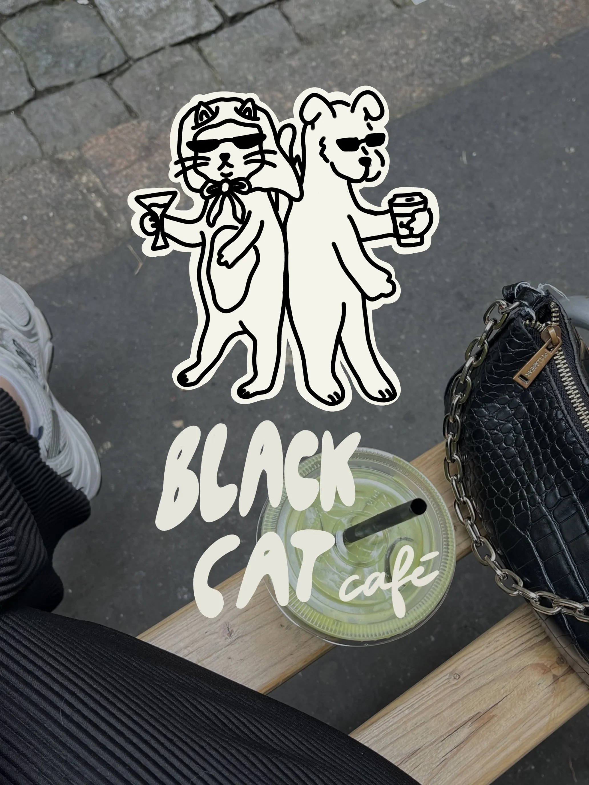 Street scene featuring a wooden table with a green beverage in a clear plastic cup with a black straw, a black quilted purse with gold chain strap, and a person wearing white sneakers and black pants, with a sticker graphic of a cat and a dog wearing sunglasses and holding drinks, over the image.