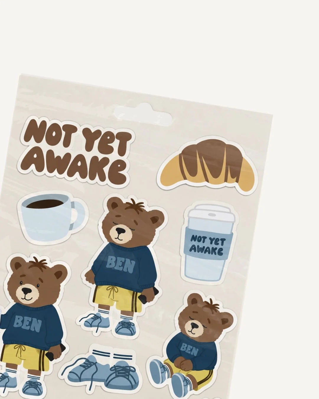 Sheet of stickers with bears, a croissant, coffee cup, and to-go coffee cup, all with the phrase "Not Yet Awake." The bears are wearing sweaters with "Ben" on them and yellow shorts, depicted in various poses.